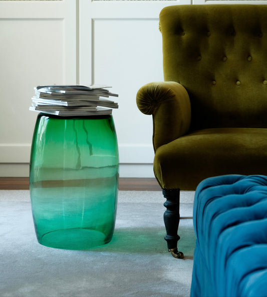 Tavolino Smeralda 50 used as a sculptural side table in emerald Murano glass, styled with stacked books beside an armchair in a contemporary interior.